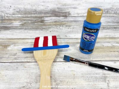 Wooden spoon with red and white hat stripes and blue painted craft stick glued on as the hat brim.