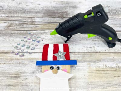 Finished Uncle Sam wooden spoon craft with red and white striped hat, blue brim, silver glitter star with rhinestone, and glue gun in the background.