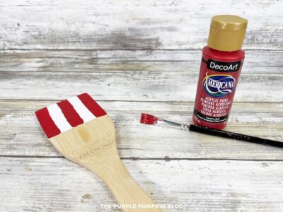 Wooden spoon with white base and red vertical stripes painted on top section to create Uncle Sam’s hat.
