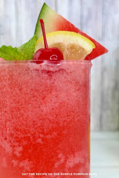 A tall glass of frozen watermelon lemonade slushie with watermelon wedge, lemon slice, cherry, and mint garnish