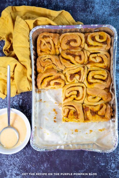 Overhead shot of a tray of pumpkin cinnamon rolls with several removed. A bowl of pumpkin glaze is on the side.
