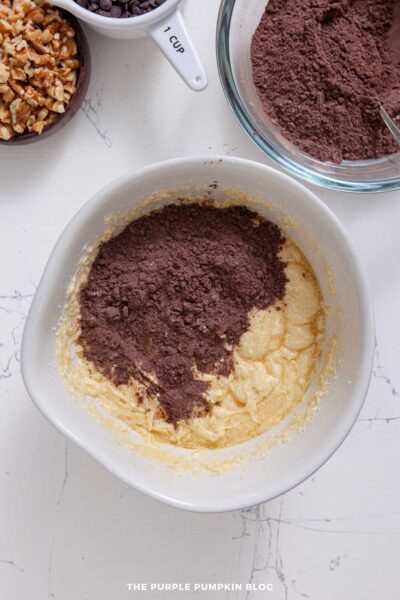 Dry cocoa mixture gradually added to wet butter and egg mixture in a white bowl. The glass bowl of remaining cocoa mixture to the side.