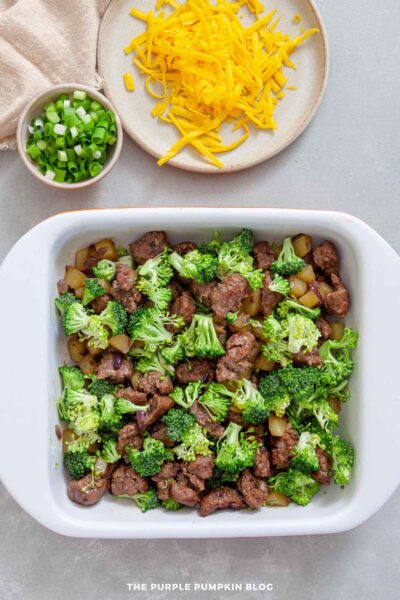 White casserole dish with a layer of browned sausage, golden potatoes, and fresh broccoli florets, ready to be topped with cheese.