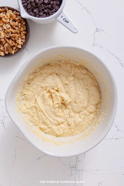 Creamed butter, sugar, eggs, vanilla mixture in a white mixing bowl, with cups of chopped walnuts and chocolate chips to the side.