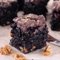 Close-up of a square chocolate walnut brownie topped with thick chocolate frosting and sprinkled with chopped nuts, with whole walnuts scattered around on the surface.