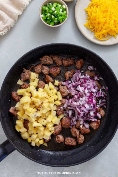 Black skillet with browned sausage pieces, raw diced potatoes, and freshly chopped red onion, sprinkled with salt and pepper, ready to cook.