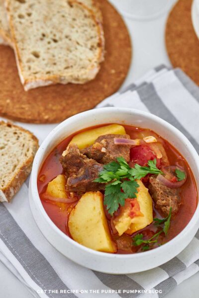 Bowl of Portuguese beef stew with beef chunks, potatoes, tomatoes, and parsley garnish, served alongside slices of rustic bread. Text overlay says: “Get the recipe on The Purple Pumpkin Blog”.
