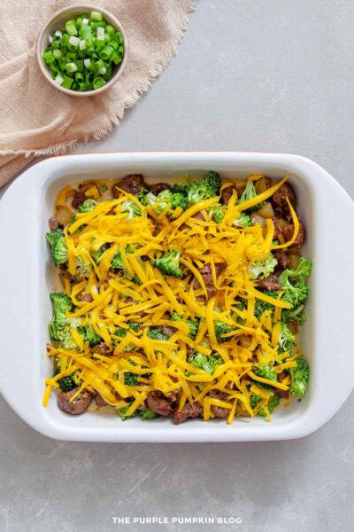 White casserole dish filled with cooked sausage, diced potatoes, and broccoli, topped generously with shredded cheddar cheese, with a bowl of chopped green onions beside it.