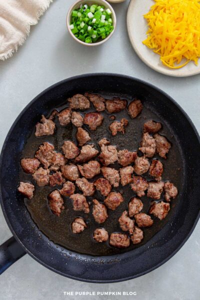 Black skillet with browned chunks of sausage cooking in their juices, with bowls of shredded cheddar cheese and chopped green onions nearby.