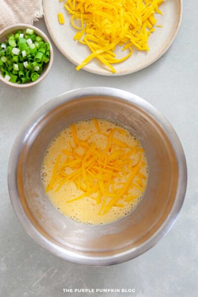 Stainless steel mixing bowl with beaten eggs and shredded cheddar cheese, with extra grated cheese and chopped green onions in bowls nearby.