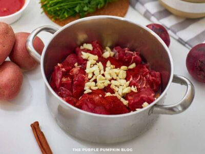 Large pot filled with raw beef cubes topped with chopped garlic and paprika, surrounded by red potatoes, parsley, onions, and a cinnamon stick.