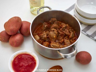 Partially cooked beef stew in a pot on a white surface, surrounded by potatoes, diced tomatoes, and stacked bowls.
