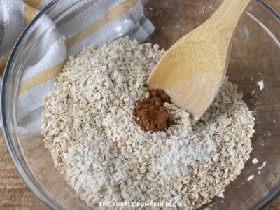 Mixing bowl with oats, flour, and pumpkin pie spice, with a wooden spoon resting on top.