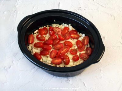 Slow cooker pot with soaked white beans topped with chopped onion and halved cherry tomatoes, ready for cooking smoky beans with bacon.