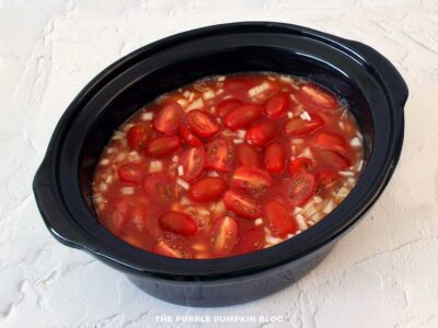 Slow cooker pot filled with white beans, chopped onion, halved cherry tomatoes, and sauce mixture poured over the top, ready to cook smoky beans with bacon.