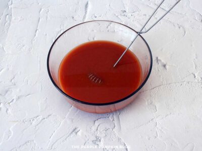 Glass mixing bowl with a whisk in smooth reddish-brown sauce made from tomato paste, BBQ sauce, molasses, vinegar, mustard, and sugar for smoky beans with bacon.