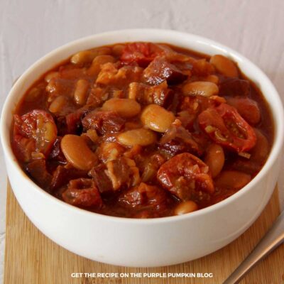 A close up image of a hearty portion of smoky beans with bacon served in a white ceramic bowl.