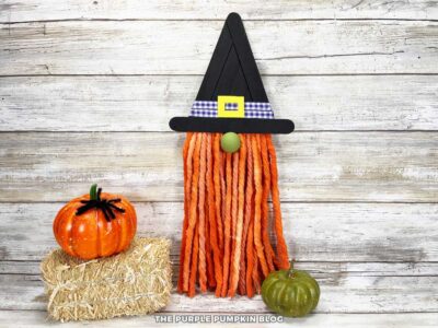 Handmade Halloween witch gnome craft made from orange mop strings for the beard, a green wooden bead for the nose, and a black craft stick witch hat decorated with a purple gingham ribbon and yellow foam buckle. The gnome is displayed on a rustic wooden background with a small green pumpkin and hay bale nearby.