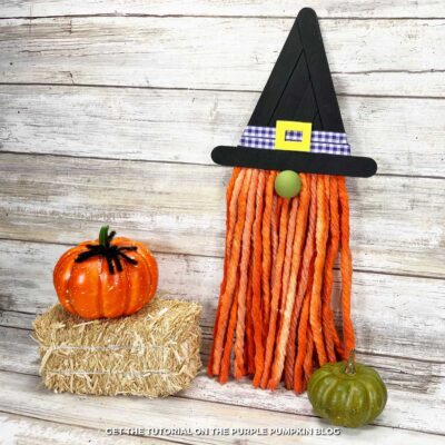 Handmade Halloween witch gnome craft made from orange mop strings for the beard, a green wooden bead for the nose, and a black craft stick witch hat decorated with a purple gingham ribbon and yellow foam buckle. The gnome is displayed on a rustic wooden background with a small green pumpkin and hay bale nearby.