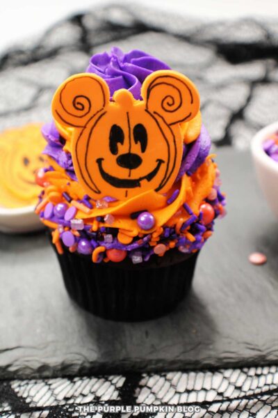 Purple Halloween cupcake with Mickey Mouse decorated chocolate topper, sprinkles, and festive orange and purple frosting.