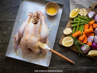 A raw turkey sits on a metal baking sheet while being brushed with melted butter using a pastry brush. A small bowl of melted butter is placed near the top of the tray. To the right, a wooden board holds chopped vegetables including carrots, celery, red onions, halved garlic bulbs, lemon wedges, and a sprig of rosemary.