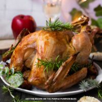 A whole roasted turkey sits on a white platter, garnished with fresh rosemary sprigs and surrounded by holiday greenery. An apple, a glass of white wine, and soft festive lights appear blurred in the background.