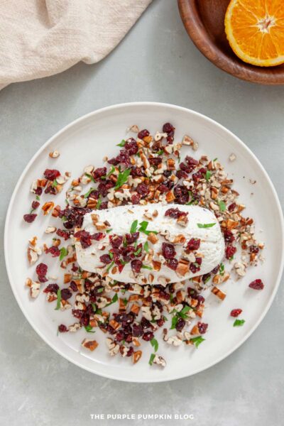 Overhead view of a partially coated cheese log placed on a white plate. Chopped dried cranberries, pecans, and parsley are scattered around and pressed onto the surface of the log. A light-colored cloth and a wooden plate with a halved orange are visible at the top of the image.
