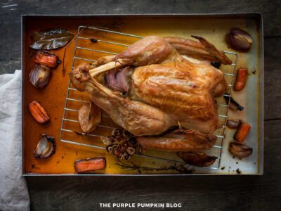 A fully roasted turkey sits on a metal rack inside a roasting pan. The turkey is golden brown with crisp skin, and roasted vegetables—including carrots, red onion pieces, bay leaves, and a halved garlic bulb—are scattered around the pan. The roasting juices coat the bottom of the pan.