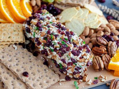 A close-up of an orange cranberry pecan cheese log coated in chopped dried cranberries, pecans, and parsley. It sits on a wooden board surrounded by assorted crackers, whole almonds, pecans, and slices of fresh oranges.