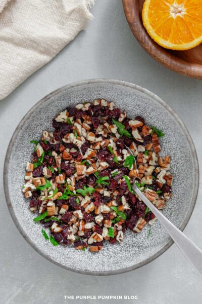 Overhead view of a bowl filled with a mixture of chopped dried cranberries, pecans, and fresh parsley. A metal spoon rests inside the bowl. Part of a wooden plate holding a halved orange and a light-colored cloth are visible at the top of the image on a gray surface.
