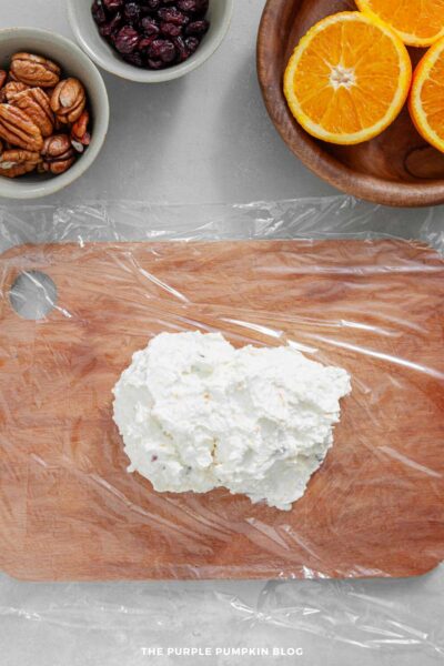 Overhead view of a wooden cutting board covered with a sheet of plastic wrap. A mound of the cream cheese mixture sits in the center, ready to be shaped. Small bowls of pecans and dried cranberries, along with a wooden plate holding halved oranges, are visible at the top of the image.