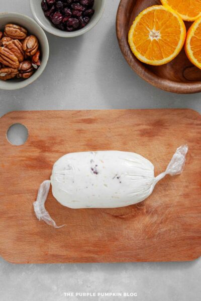 Overhead view of a cream cheese mixture wrapped tightly in plastic wrap and shaped into a log. It sits on a wooden cutting board. Small bowls of pecans and dried cranberries, along with a wooden plate holding halved oranges, are visible at the top of the image.