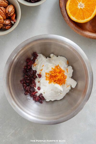 Overhead view of a metal mixing bowl containing cream cheese, a pile of dried cranberries, and a mound of orange zest. Surrounding the bowl are small dishes of pecans and cranberries, along with a wooden plate holding half an orange on a light gray surface.