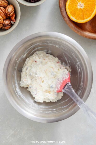 Overhead view of a metal mixing bowl filled with a blended cream cheese mixture containing small bits of cranberries and orange zest. A red silicone spatula rests in the bowl. Small bowls of pecans and cranberries, along with half an orange on a wooden plate, are visible at the top of the image.