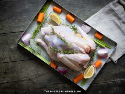 A raw whole turkey sits on a metal roasting rack inside a roasting pan, topped with fresh rosemary sprigs. Chopped vegetables including carrots, celery, and red onion pieces are arranged around the turkey along with bay leaves and lemon wedges. A beige kitchen towel rests beside the pan on a dark rustic wooden surface.