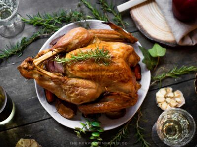 A whole roasted turkey sits on a white platter, topped with fresh rosemary sprigs and surrounded by roasted vegetables. The platter is set on a rustic wooden table decorated with greenery, cut garlic bulbs, a glass of white wine, and a wooden serving board.