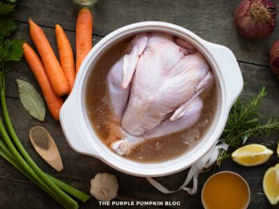 A whole raw turkey sits submerged in a brine inside a large white pot. Around the pot are ingredients including whole carrots, celery stalks, bay leaves, a halved garlic bulb, lemon wedges, red onions, rosemary, black pepper, and a bowl of melted butter. All items are arranged on a dark rustic wooden surface.