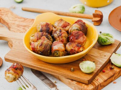 Maple glazed bacon wrapped Brussels sprouts served in a yellow baking dish on a wooden board, with whole and halved Brussels sprouts scattered around.