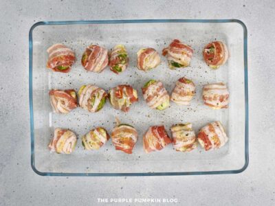 Bacon wrapped Brussels sprouts arranged in rows inside a clear glass baking dish, seasoned and ready for baking.