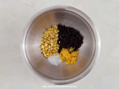 Chopped pistachios, chopped walnuts, dried blackcurrants, lemon zest, and salt in a metal mixing bowl.