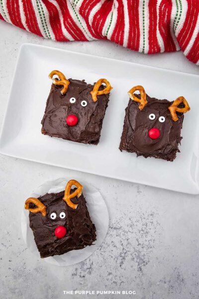 Three reindeer brownies decorated with chocolate frosting, pretzel antlers, candy eyes, and red M&M noses arranged on a white serving plate and parchment paper.
