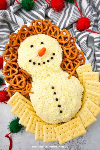 Snowman-shaped cheeseball coated in shredded white cheddar cheese with peppercorn eyes, mouth, and buttons and a baby carrot nose, served on a round platter with pretzels and crackers for a Christmas appetizer.