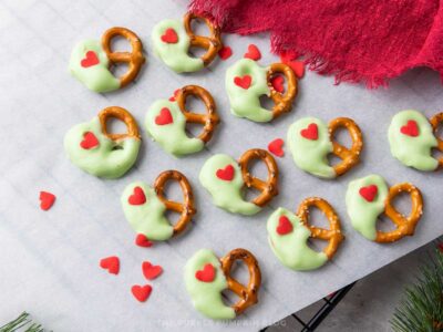 Green candy-coated pretzel twists decorated with red heart sprinkles arranged on a gray surface, with scattered heart sprinkles around them.