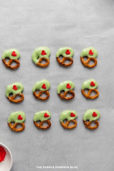 Green candy-coated pretzel twists decorated with red heart sprinkles arranged in neat rows on parchment paper.
