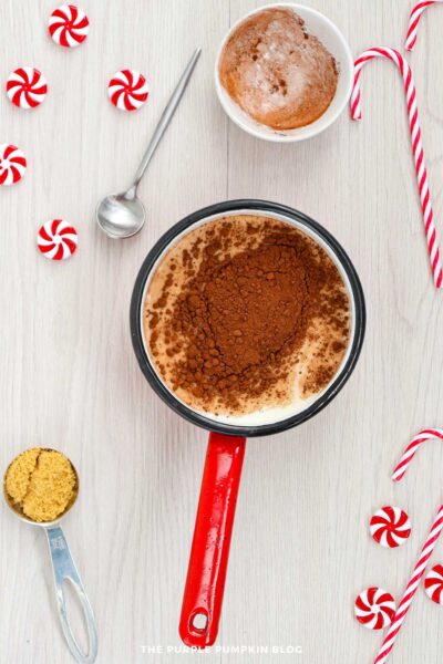 A saucepan of warm milk topped with a mound of cocoa powder, with a bowl of used cocoa powder, peppermint candies, candy canes, and a measuring spoon of brown sugar arranged around it on a light wooden surface.