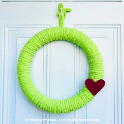 Green yarn Grinch wreath hanging on a white front door with a red felt heart accent attached to the side.
