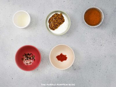 Small bowls containing maple syrup, whole grain mustard, spices, and seasoning arranged on a gray surface.