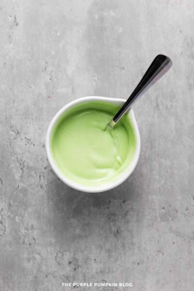 A white bowl filled with smooth melted green candy melts, with a spoon resting inside on a gray surface.