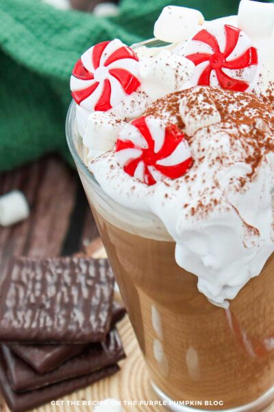 Close-up of whipped cream piled high on a glass of peppermint hot chocolate, decorated with cocoa powder, mini marshmallows, and red-and-white peppermint candies. Chocolate mint cookies sit beside the glass on a wooden slice, with a green knitted blanket in the background.
