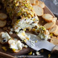 Pistachio and blackcurrant cheese log coated in chopped pistachios and walnuts on a wooden board with sliced crackers and a cheese knife spreading the cheese.